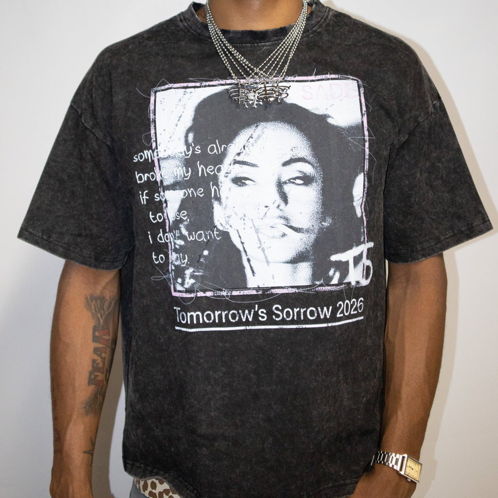 A man wearing a faded black graphic t-shirt with a portrait of Sade and the text 'Tomorrow's Sorrow 2026', paired with a camouflage baseball cap that reads 'GOD'S FAVORITE'.