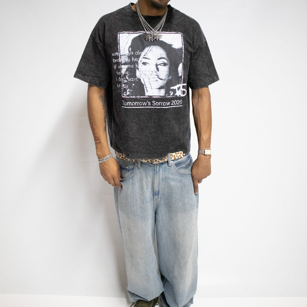 A man wearing a black graphic t-shirt with a Sade print, light wash baggy jeans, a camouflage baseball cap, and green sneakers, standing against a white studio background.